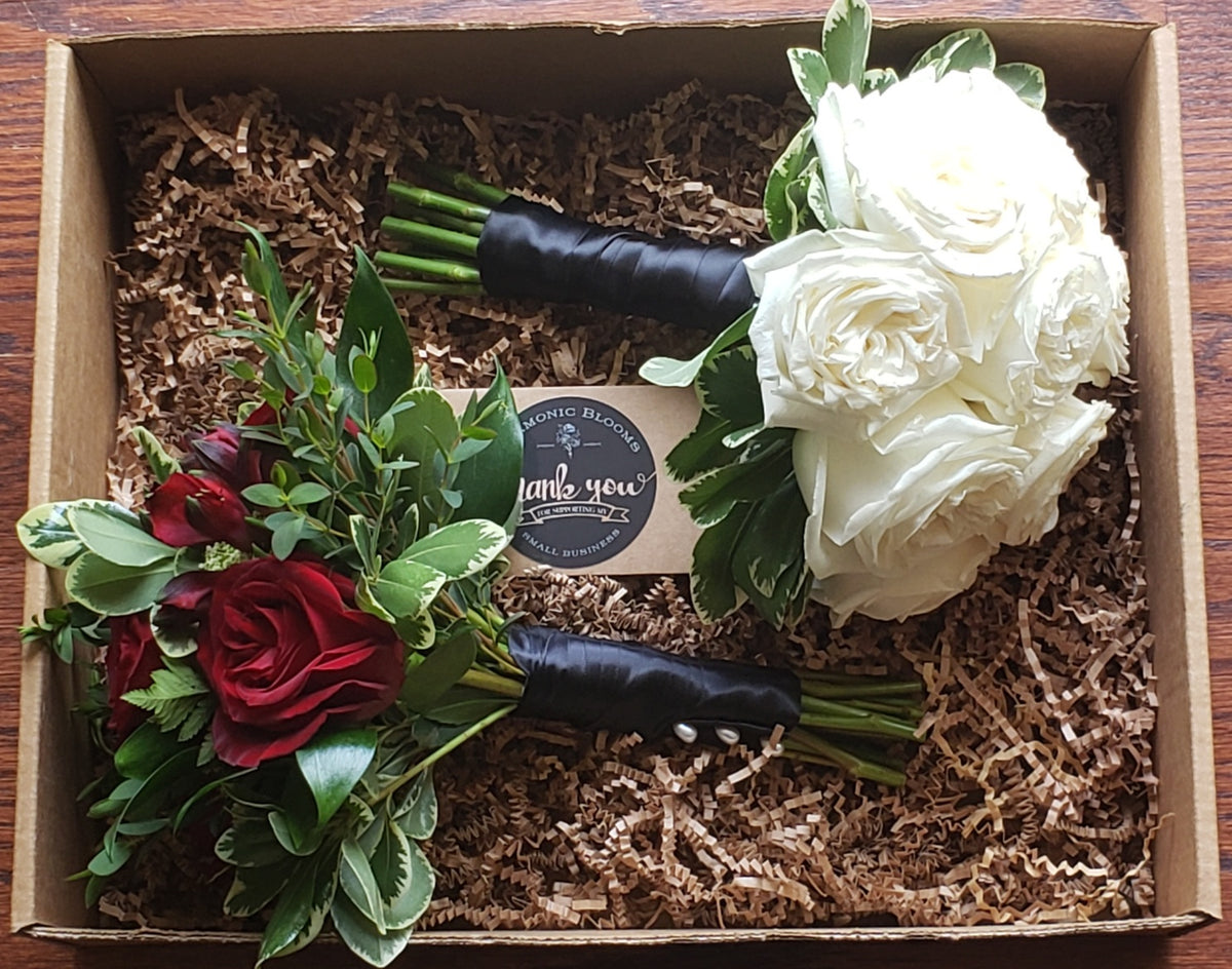 A floral gift set in a cardboard box filled with brown paper shred, featuring all white rose bouquet, a red mixed flower bouquet with green foliage,  both wrapped with a black satin ribbon with a round Harmonic Blooms logo tag placed in between them that reads 'thank you'.