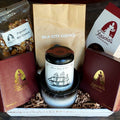 A curated gift basket on a dark wooden surface, featuring a Silk City Coffee, a jar of Oliver Pluff & Co tea, a bag of Fascia's Popcorn, and Fascia's Chocolates in its original packaging, all arranged in a white wooden box with shredded paper filler by Harmonic Blooms in Glastonbury, CT.