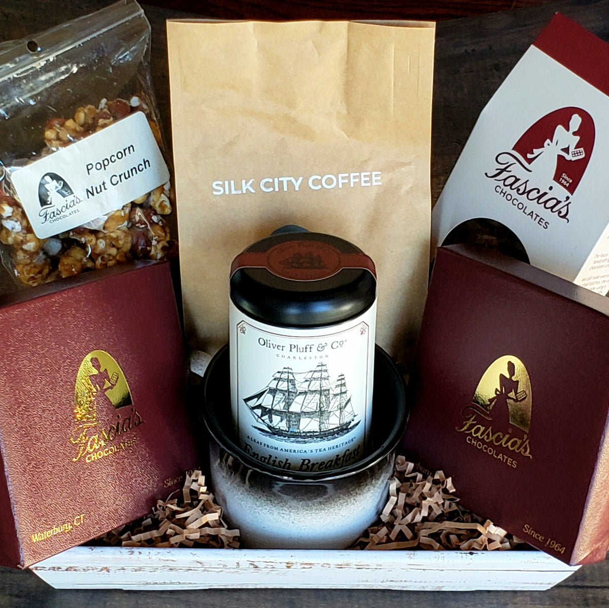 A curated gift basket on a dark wooden surface, featuring a Silk City Coffee, a jar of Oliver Pluff & Co tea, a bag of Fascia's Popcorn, and Fascia's Chocolates in its original packaging, all arranged in a white wooden box with shredded paper filler by Harmonic Blooms in Glastonbury, CT.