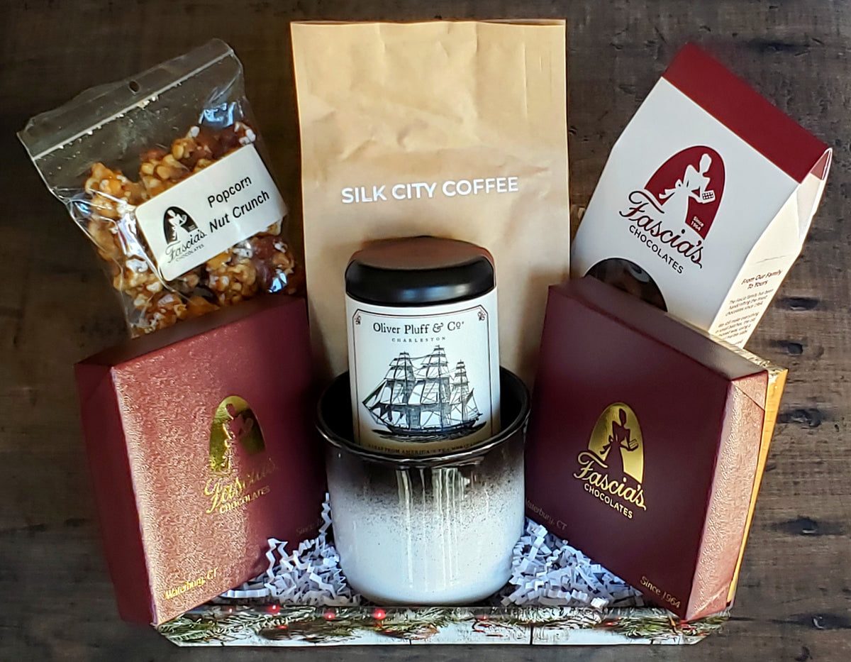 A curated gift basket on a dark wooden surface, featuring a Silk City Coffee, a jar of Oliver Pluff & Co tea, a bag of Fascia's Popcorn, and Fascia's Chocolates in its original packaging, all arranged in a white wooden box with shredded paper filler by Harmonic Blooms in Glastonbury, CT.
