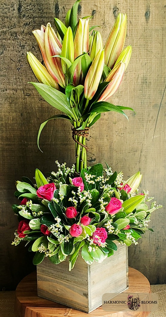 A topiary floral arrangement featuring a cluster of tall pink lilies and bright pink spray roses with green foliage in a square wooden box, designed by Harmonic Blooms in Glastonbury, CT and displayed on a wooden pedestal against a rustic wood wall.