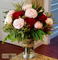 A vibrant floral arrangement of red and light pink roses, peonies, ranunculus and green foliage in a clear glass vase with a burlap-wrapped neck, designed by Harmonic Blooms in Glastonbury, CT and set on a granite countertop in front of a white door.