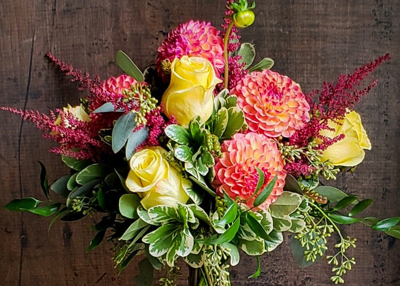 A vibrant floral bouquet featuring yellow roses, pink and coral dahlias, and green foliage, wrapped in burlap and displayed in a glass vase on a wooden surface against a dark wood plank background.