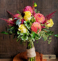 A vibrant floral bouquet featuring yellow roses, pink and coral dahlias, and green foliage, wrapped in burlap designed by Harmonic Blooms in Glastonbury, CT and displayed in a glass vase on a wooden surface against a dark wood plank background.