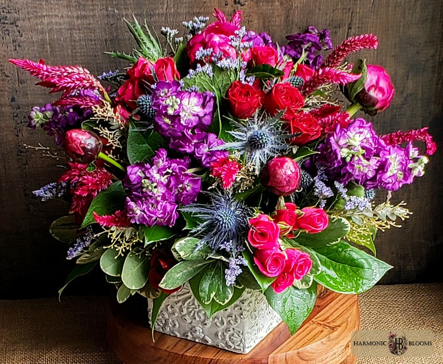 A vibrant floral arrangement featuring red and pink roses, purple stock, blue thistle, and green foliage, presented in a textured white ceramic pot on a wooden surface against a dark rustic background.
