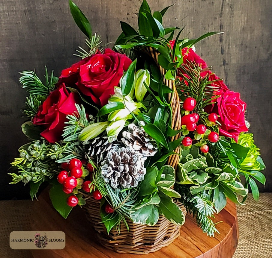 A festive holiday floral arrangement in a woven basket, featuring mums, red roses, assorted evergreens, pinecones, and red berries, designed by Harmonic Blooms in Glastonbury, CT and displayed on a wooden surface against a dark wood background.