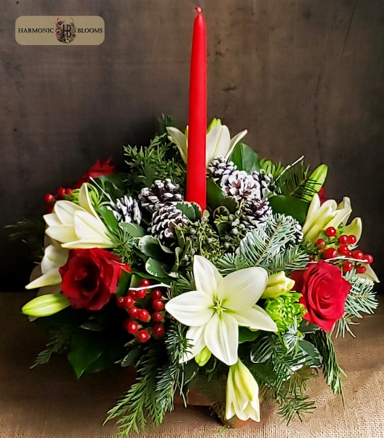 A festive Christmas floral arrangement featuring white lilies, red roses, green pine branches, pinecones, red hypericum, and a single tall red taper candle, displayed on a rustic brown surface against a dark wood background. A 'Harmonic Blooms' logo is visible in the top left corner.