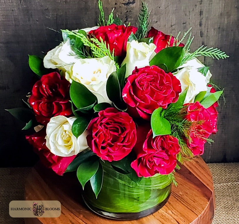 A vibrant floral arrangement featuring red and white roses with green foliage, designed by Harmonic Blooms in Glastonbury, CT and presented in a glass vase on a wooden surface against a dark rustic background.