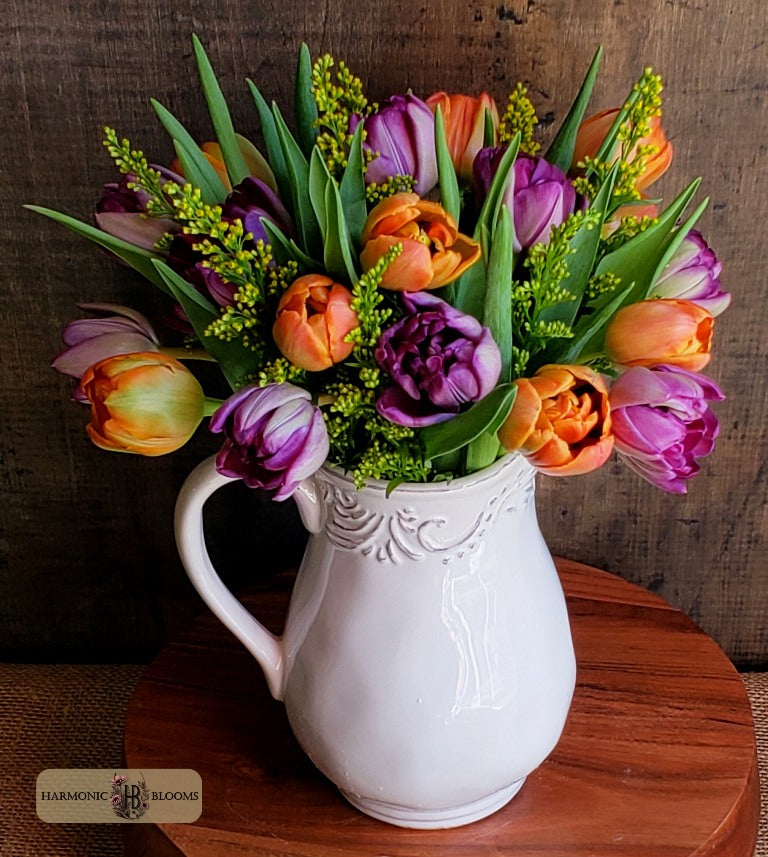A vibrant tulip bouquet in a white ceramic pitcher vase, set on a round wooden table against a dark rustic wood background. The bouquet features orange and purple tulips with green foliage designed by Harmonic Blooms in Glastonbury, CT.