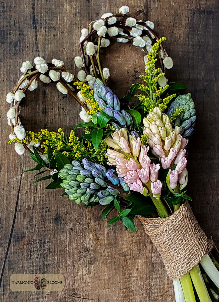 A rustic heart-shaped floral arrangement made from pussy willow with pastel pink and lavender hyacinth, green leaves, yellow solidago,  wrapped in burlap, designed by Harmonic Blooms in Glastonbury, CT, and set on a dark wooden surface. A small 'Harmonic Blooms' logo is visible in the bottom-left corner.