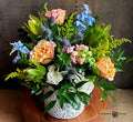 A vibrant floral arrangement featuring peach roses, blue delphiniums, thistles, and green foliage in a white textured ceramic pot designed by Harmonic Blooms in Glastonbury, CT and set on a wooden surface against a dark rustic background.
