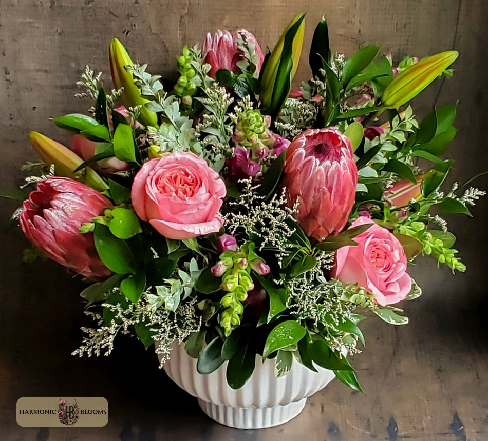 A vibrant floral arrangement featuring pink protea roses, pink roses, snapdragons, green foliage, and pink lilies in a white ribbed ceramic pot, designed by Harmonic Blooms in Glastonbury, CT and set against a dark rustic wooden background. A 'Harmonic Blooms' logo is visible in the bottom-left corner.