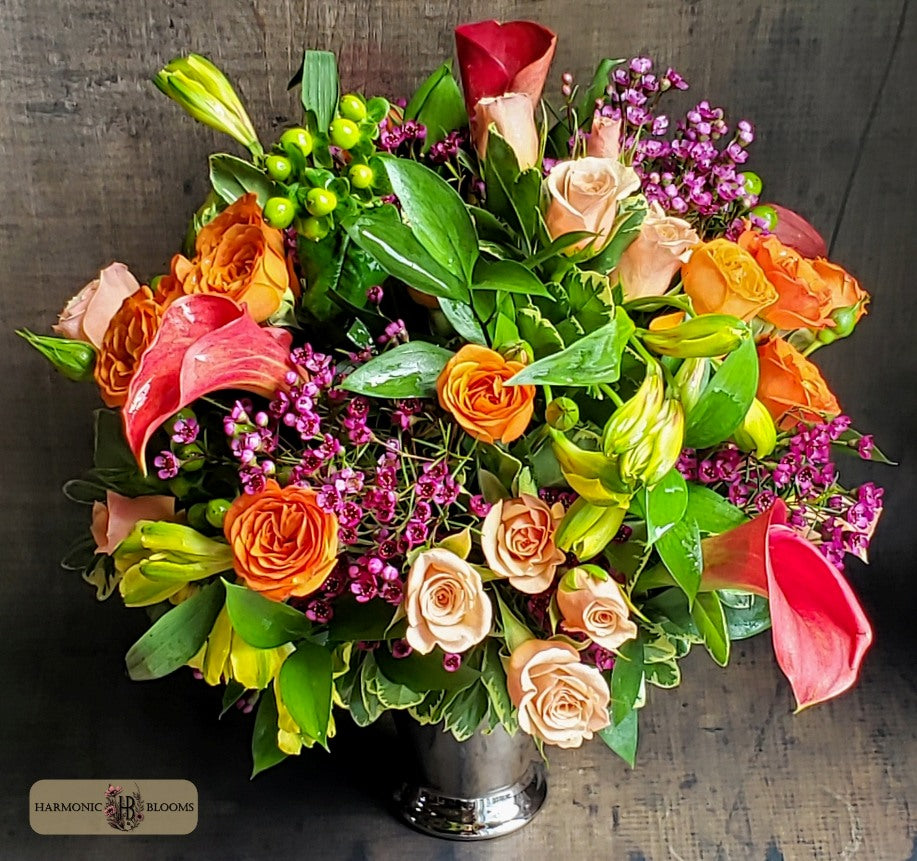 A vibrant artificial flower bouquet in a silver metal vase, featuring orange roses, peach calla lilies, red tulips, green foliage, and purple filler flowers, designed by Harmonic Blooms in Glastonbury, CT and set against a dark rustic wooden background.