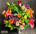 A vibrant artificial flower bouquet in a silver metal vase, featuring orange roses, peach calla lilies, red tulips, green foliage, and purple filler flowers, designed by Harmonic Blooms in Glastonbury, CT and set against a dark rustic wooden background.