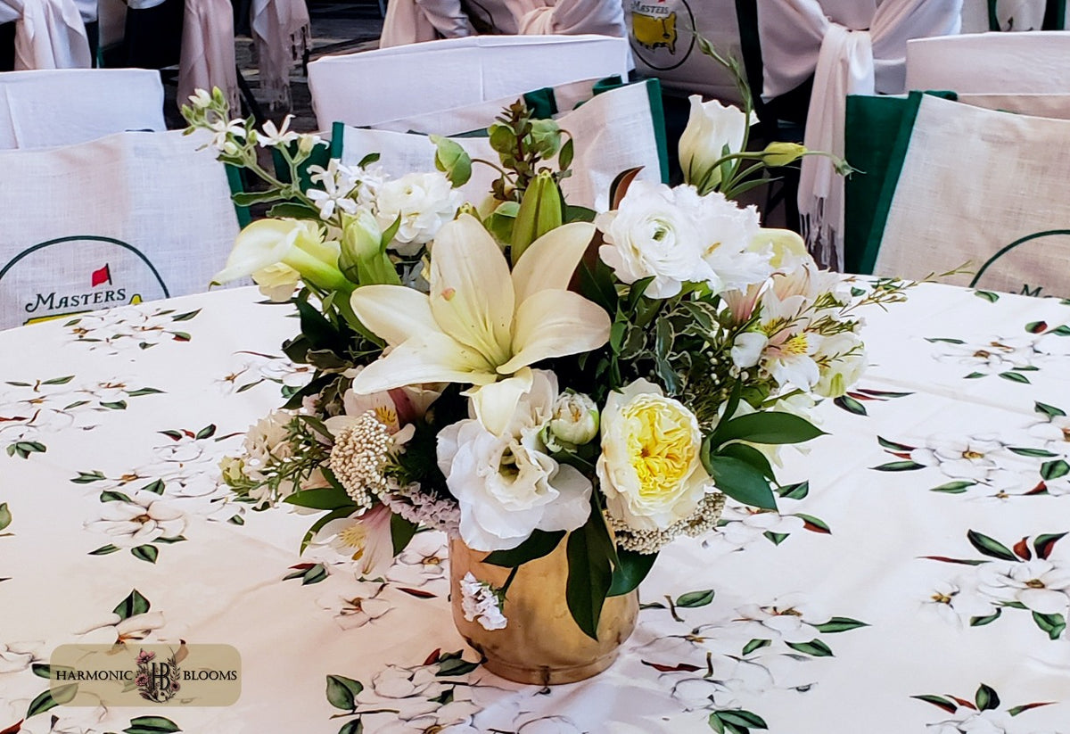 A floral centerpiece arrangement in a gold vase on a decorated tablecloth, featuring white lilies, white roses, and green foliage, with chairs and event decor blurred in the background designed for the First Lady's Luncheon in Washington D.C. by Harmonic Blooms in Glastonbury, CT.