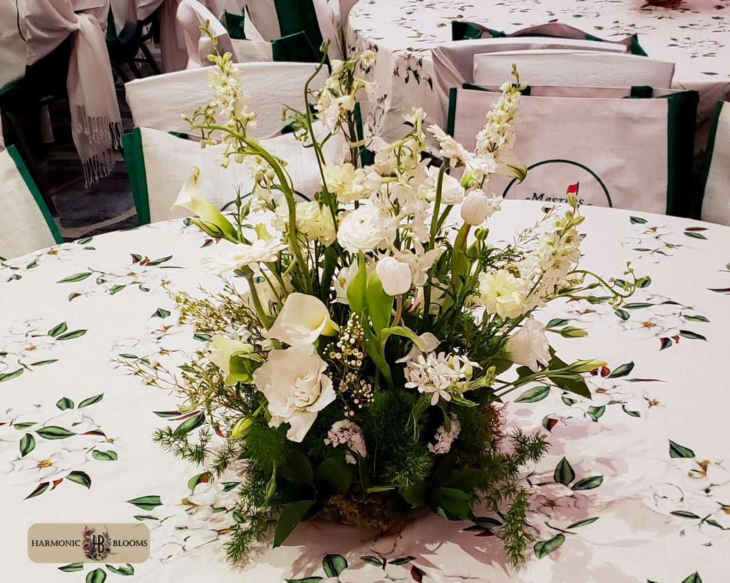 A floral centerpiece arrangement of white flowers and green foliage, designed by Harmonic Blooms in Glastonbury, CT and placed on a round table with a white tablecloth printed with delicate green leaves. In the background, white chairs with green trim and branded bags are visible.