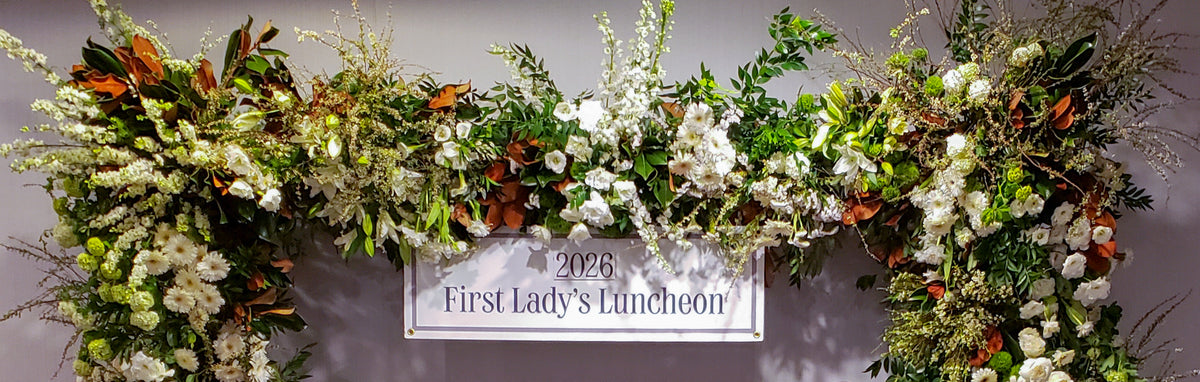 A large floral arch arrangement with white flowers, green foliage, and brown leaves, with a central sign reading '2026 First Lady's Luncheon' in Wasington DC and designed by Harmonic Blooms.