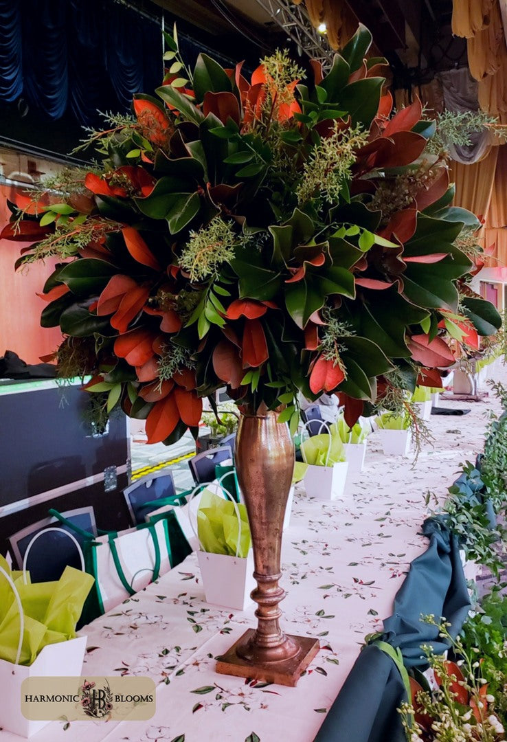 A large floral centerpiece arrangement with glossy green leaves and rust-orange flowers in a tall bronze-toned pedestal vase, displayed on a decorated event table designed for the First Lady's Luncheon in Washington D.C. by Harmonic Blooms.