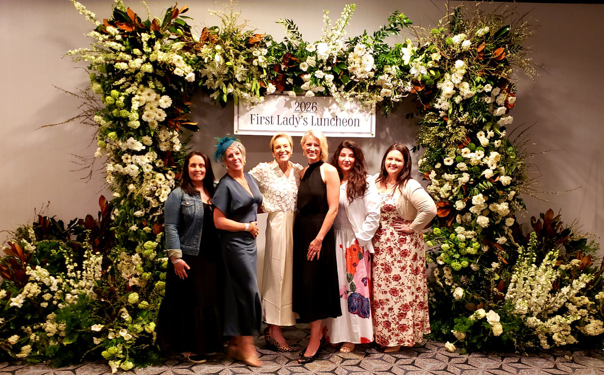 Six women posing together in front of a large floral arch backdrop with a sign reading '2025 First Lady's Luncheon'. The women are wearing formal dresses, and the background is dominated by white flowers, green foliage, and brown leaves.