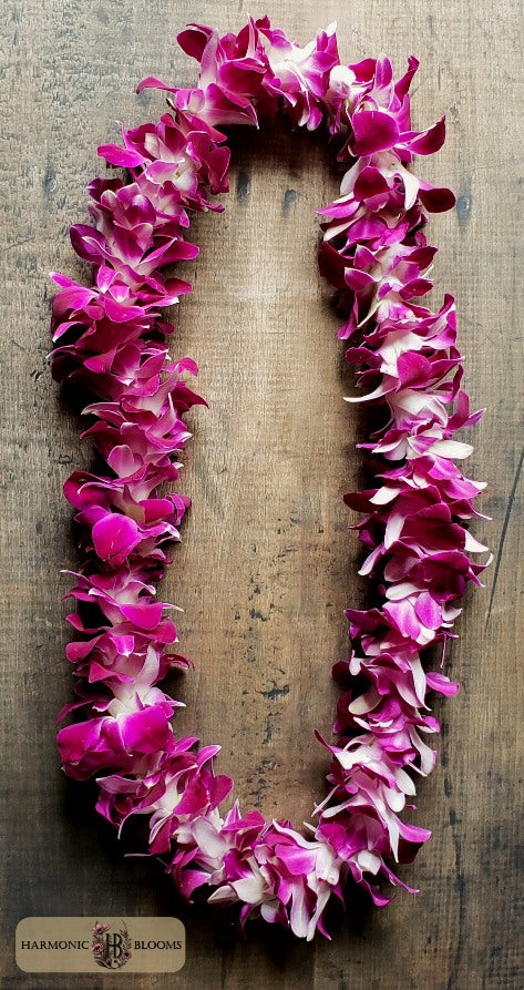 A vibrant pink and white orchid flower lei necklace arranged in an oval shape on a rustic wooden background, with a 'Harmonious Blooms' brand logo in the bottom-left corner.