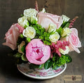 A floral arrangement featuring light pink and white roses, a large pink peony, green foliage, and pink filler flowers, presented in a floral-patterned ceramic teacup, designed by Harmonic Blooms in Glastonbury, CT and set against a dark rustic wooden background. A 'Harmonic Blooms' logo is visible in the bottom right corner.