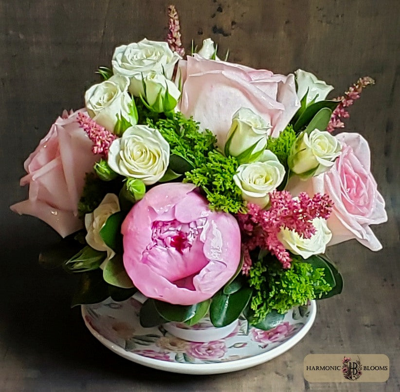 A floral arrangement featuring light pink and white roses, a large pink peony, green foliage, and pink filler flowers, presented in a floral-patterned ceramic teacup, designed by Harmonic Blooms in Glastonbury, CT and set against a dark rustic wooden background. A 'Harmonic Blooms' logo is visible in the bottom right corner.