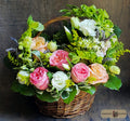 A wicker flower basket filled with a colorful bouquet of pink and peach roses, white flowers, green foliage, and small yellow-green filler stems, designed by Harmonic Blooms in Glastonbury, CT and set against a dark rustic wooden background. A 'Harmonic Blooms' logo is visible in the bottom right corner.
