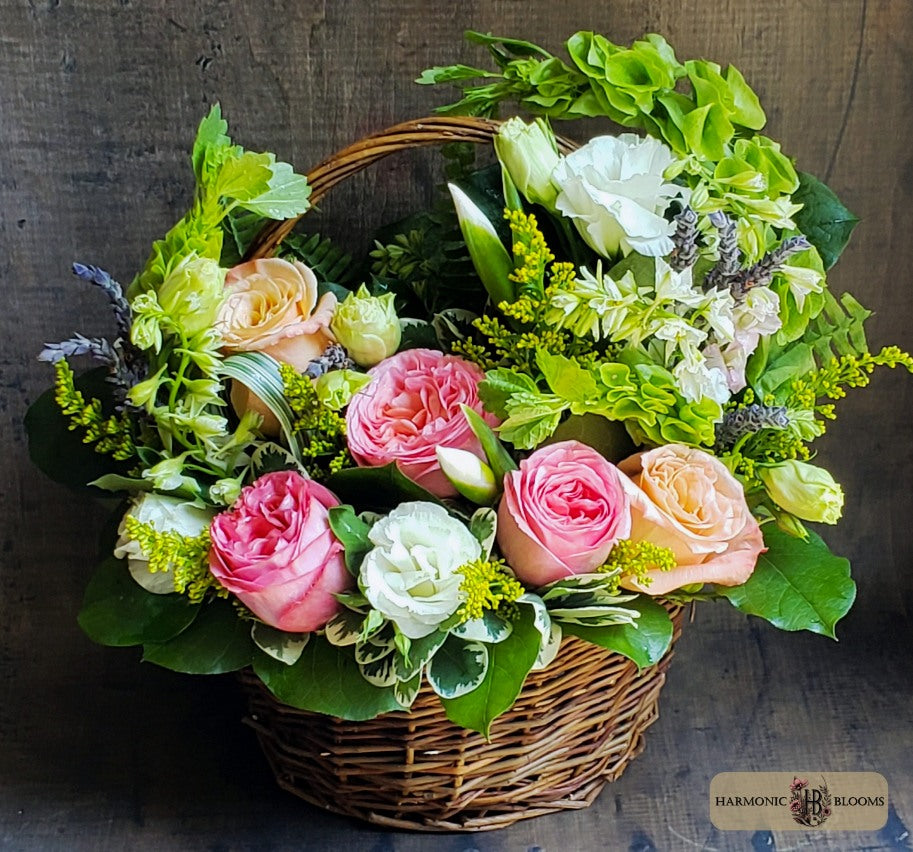 A wicker flower basket filled with a colorful bouquet of pink and peach roses, white flowers, green foliage, and small yellow-green filler stems, designed by Harmonic Blooms in Glastonbury, CT and set against a dark rustic wooden background. A 'Harmonic Blooms' logo is visible in the bottom right corner.