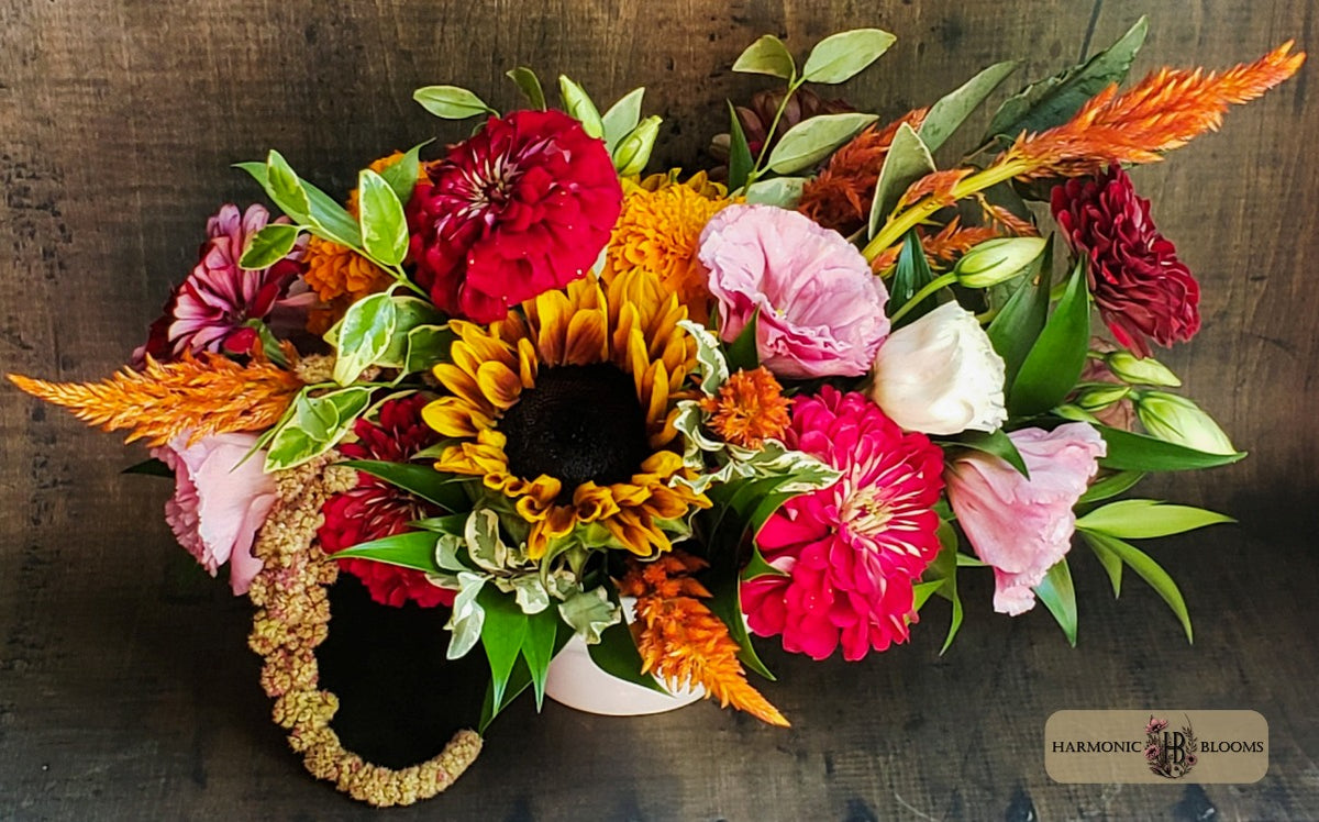 A vibrant floral arrangement featuring a yellow sunflower, red and pink zinnias, pink roses, orange feather grass, and green foliage, presented in a white vase designed by Harmonic Blooms in Glastonbury, CT and set on a dark rustic wooden background. Amaranthus is draped over the left side of the vase, and a 'Harmonic Blooms' logo is visible in the bottom right corner.