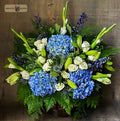 A vibrant floral arrangement featuring blue hydrangeas, white roses, white lilies, white gladiolus and blue delphiniums with green foliage, presented in a rustic wooden basket designed by Harmonic Blooms in Glastonbury, CT and placed against a dark wood background.