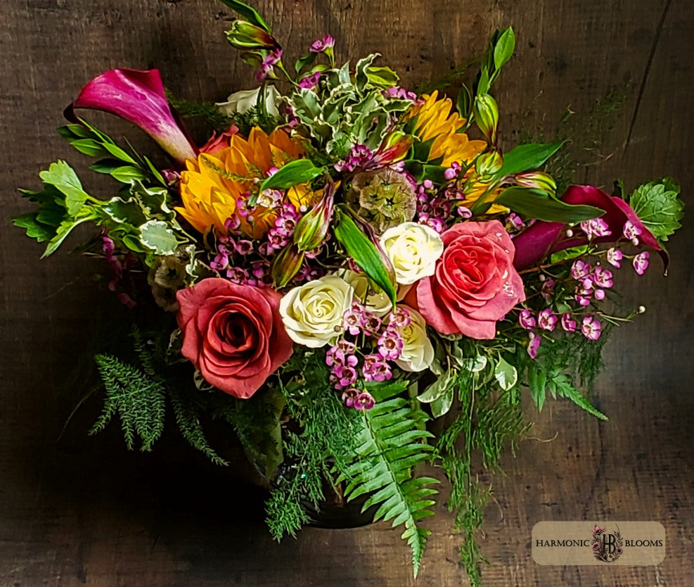 A vibrant floral arrangement featuring pink and red roses, yellow sunflowers, magenta calla lilies, green ferns, and small purple filler flowers, presented in a glass vase, designed by Harmonic Blooms in Glastonbury, CT and placed on a rustic dark wood background.
