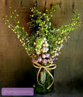 A dried floral arrangement featuring green stems, small buds, and clusters of pale pink flowers, styled in a dark green glass jar with a tan ribbon bow, set against a rustic wooden background.