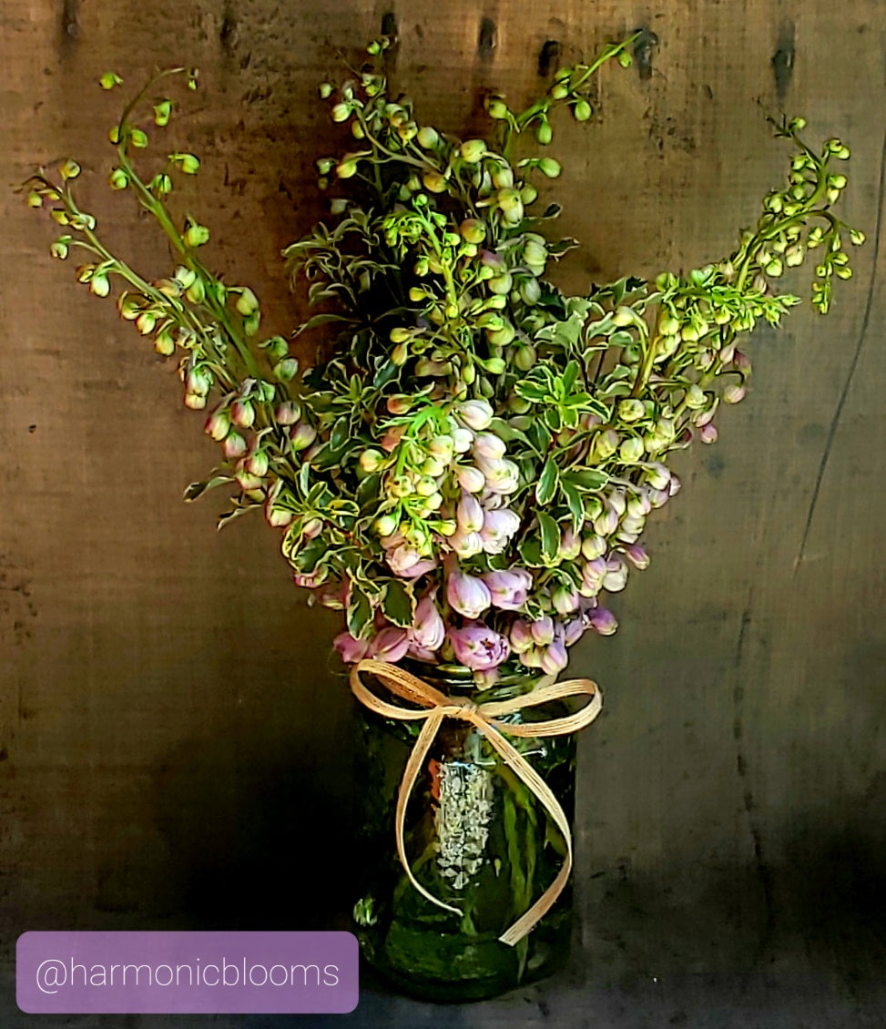 A dried floral arrangement featuring green stems, small buds, and clusters of pale pink flowers, styled in a dark green glass jar with a tan ribbon bow, set against a rustic wooden background.