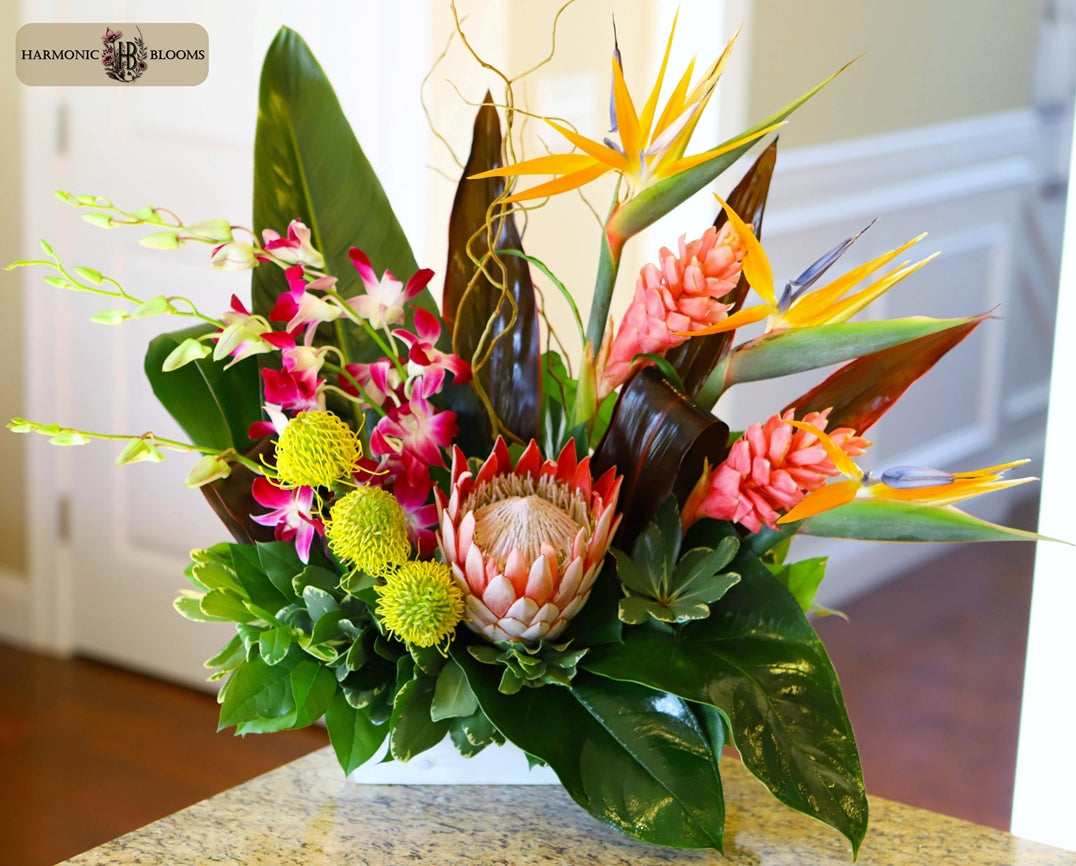 A vibrant tropical flower arrangement featuring a pink protea, yellow bird of paradise, pink and yellow orchids, and green foliage, designed by Harmonic Blooms in Glastonbury, CT and displayed in a white wooden rectangular container on a granite countertop.