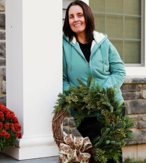 Owner of Harmonic Blooms wearing a teal zip-up hoodie and black pants stands on a stone porch holding a green evergreen wreath with a large beige bow decorated with red berries.