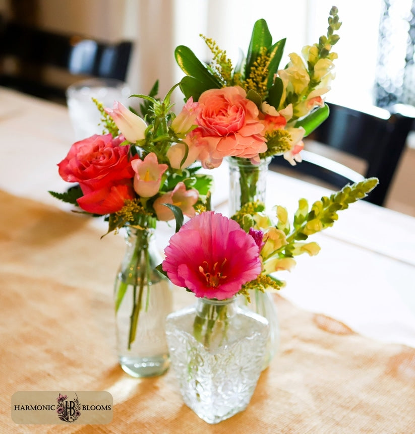Three clear glass vases filled with a colorful floral arrangement, including pink, coral, peach, and yellow blossoms, set on a beige tablecloth in a softly blurred indoor setting at 2 Hopewell Bistro & Bar in South Glastonbury, CT. A logo for Harmonic Blooms appears in the lower-left corner.
