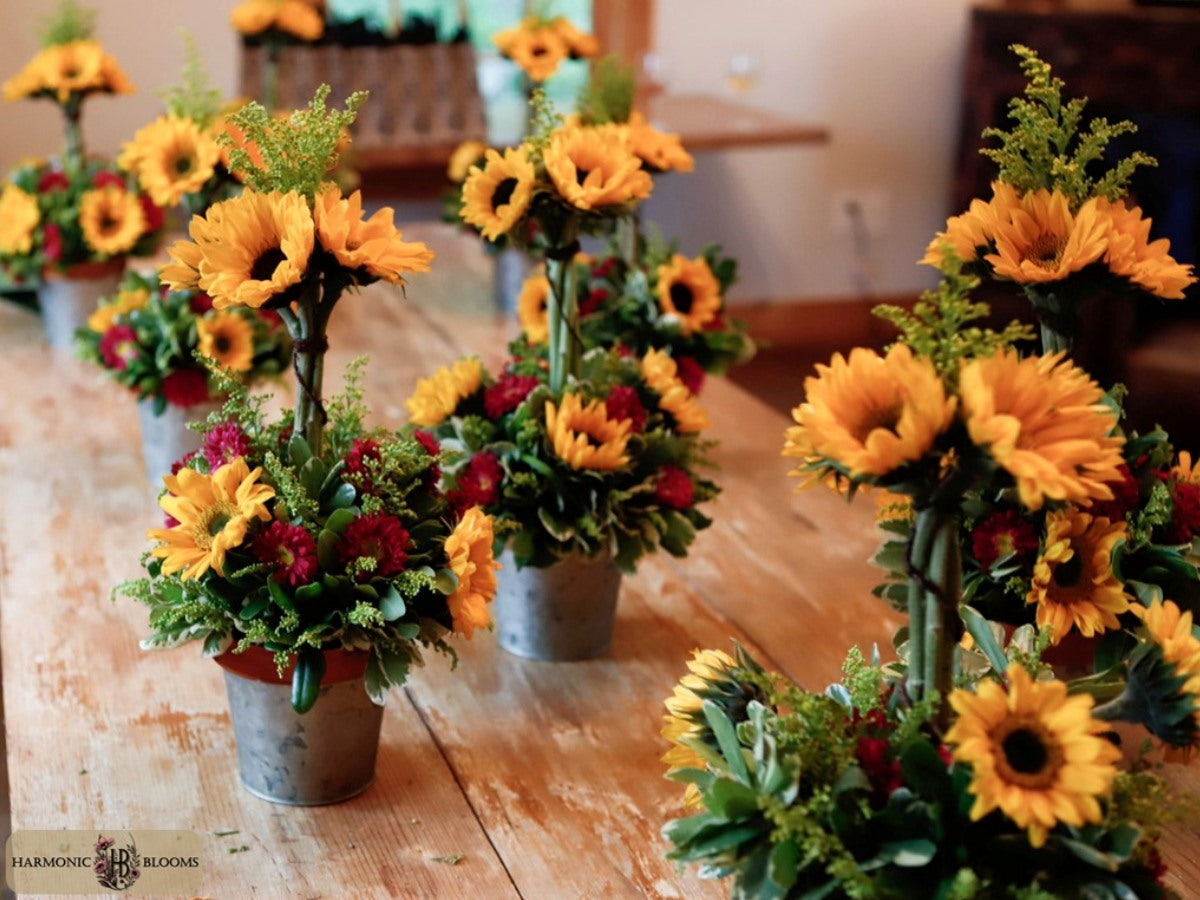 A series of small metal bucket flower arrangements for a floral workshop by Harmonic Blooms featuring bright yellow sunflowers, green foliage, and red flowers, displayed on a rustic wooden table at Crystal Ridge Winery in South Glastonbury, CT.