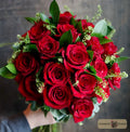 A large bouquet of vibrant red roses with green leaves and small green filler flowers designed by Harmonic Blooms in Glastonbury, CT against a dark rustic wooden background. A 'Harmonic Blooms' logo is visible in the bottom right corner.