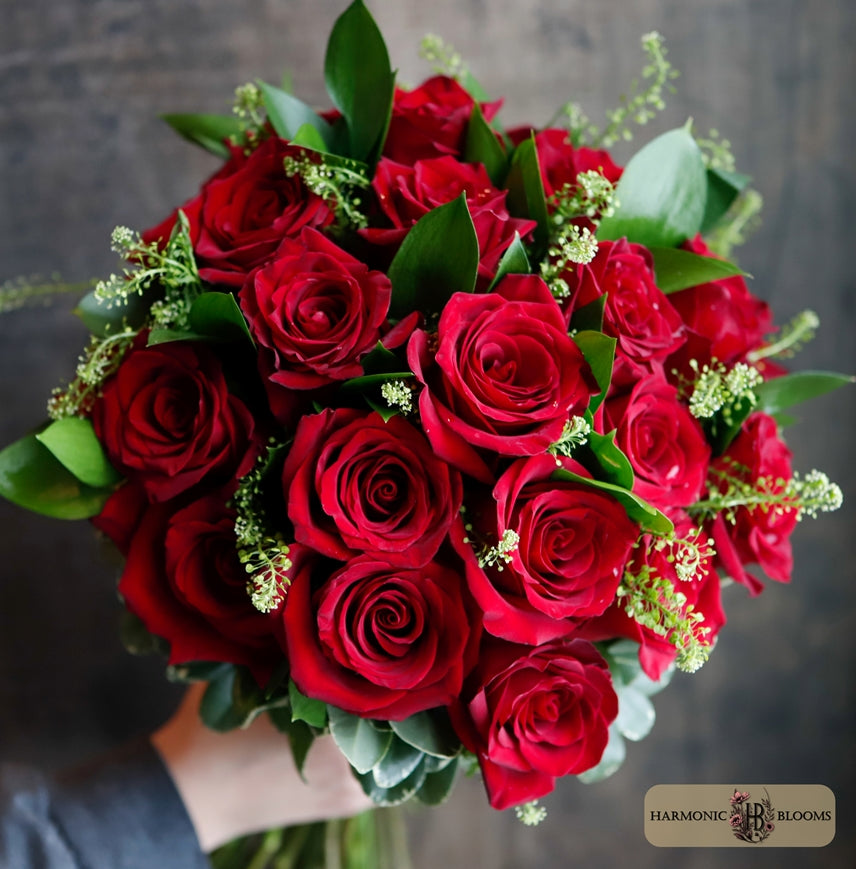 A large bouquet of vibrant red roses with green leaves and small green filler flowers designed by Harmonic Blooms in Glastonbury, CT against a dark rustic wooden background. A 'Harmonic Blooms' logo is visible in the bottom right corner.