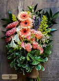 A vibrant floral bouquet featuring pink and orange gerbera daisies, peach roses, white calla lilies, purple lavender, and green foliage, wrapped in a brown jute twine designed by Harmonic Blooms in Glastonbury, CT and displayed on a dark rustic wooden background. A 'Harmony Blooms' logo is visible in the bottom-left corner.