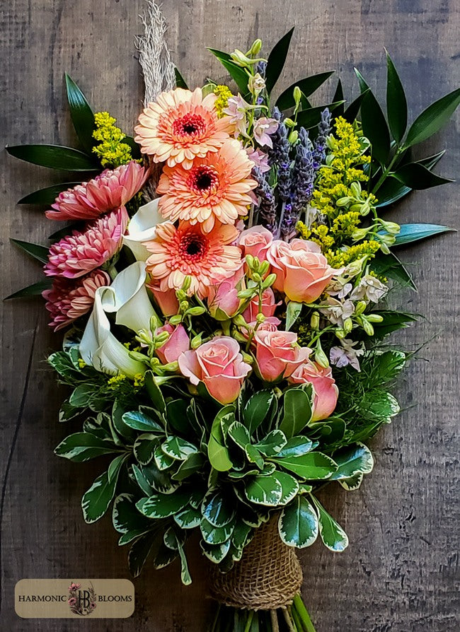 A vibrant floral bouquet featuring pink and orange gerbera daisies, peach roses, white calla lilies, purple lavender, and green foliage, wrapped in a brown jute twine designed by Harmonic Blooms in Glastonbury, CT and displayed on a dark rustic wooden background. A 'Harmony Blooms' logo is visible in the bottom-left corner.