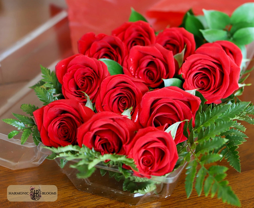 A bouquet of ten vibrant red roses with green leaves arranged in a clear plastic container, designed by Harmonic Blooms in Glastonbury, CT set on a wooden surface with a blurred red background.
