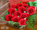 A bouquet of ten vibrant red roses with green leaves arranged in a clear plastic container, designed by Harmonic Blooms in Glastonbury, CT set on a wooden surface with a blurred red background.