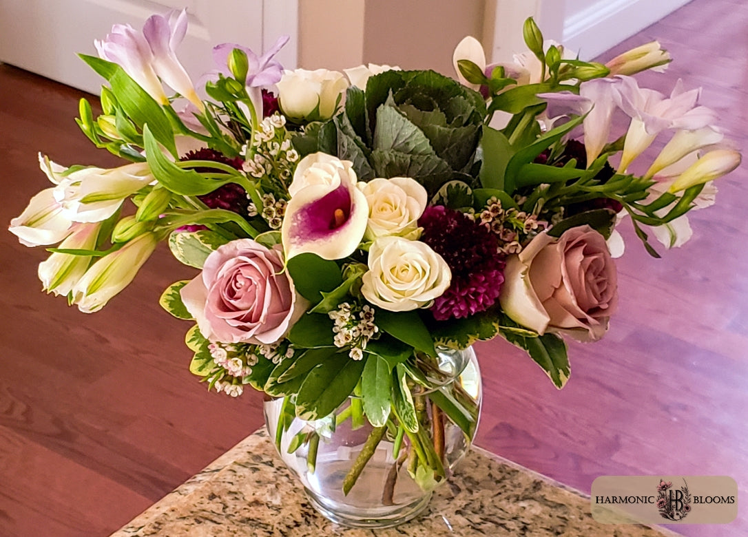 A vibrant floral arrangement in a clear glass vase, featuring pink and cream roses, white calla lilies, purple alstroemeria, and green foliage, set on a granite countertop with a wooden floor in the background by Harmonic Blooms in Glastonbury, CT.