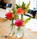 Three clear glass vases filled with a colorful floral arrangement, including pink, coral, peach, and yellow blossoms, set on a beige tablecloth in a softly blurred indoor setting. A logo for Harmonic Blooms appears in the lower-left corner.