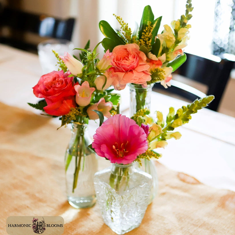 Three clear glass vases filled with a colorful floral arrangement, including pink, coral, peach, and yellow blossoms, set on a beige tablecloth in a softly blurred indoor setting. A logo for Harmonic Blooms appears in the lower-left corner.