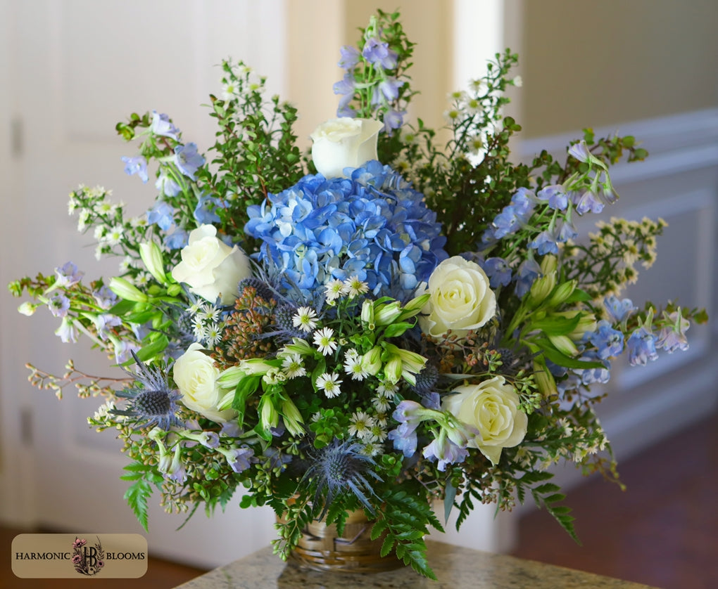 A vibrant floral arrangement featuring blue hydrangeas, white roses, blue delphiniums, and green foliage in a wicker basket designed by Harmonic Blooms in Glastonbury, CT and set on a tabletop with a blurred indoor background.