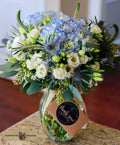 A vibrant floral bouquet in a clear glass jar vase, featuring light blue hydrangeas, white roses, green foliage, and blue thistles, tied with a brown paper tag that reads 'thank you' designed by Harmonic Blooms in Glastonbury, CT.