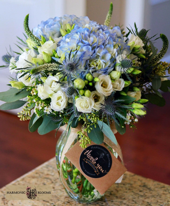 A vibrant floral bouquet in a clear glass jar vase, featuring light blue hydrangeas, white roses, green foliage, and blue thistles, tied with a brown paper tag that reads 'thank you' designed by Harmonic Blooms in Glastonbury, CT.