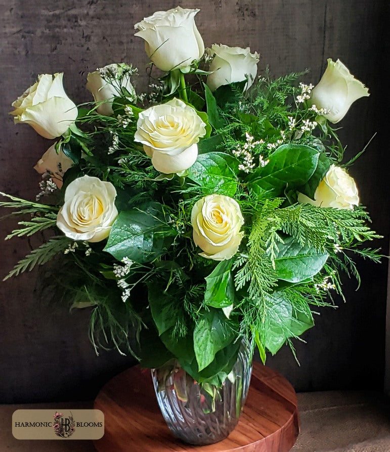 A floral arrangement of white roses and green foliage in a clear glass vase, designed by Harmonic Blooms in Glastonbury, CT and displayed on a round wooden surface against a dark textured background. A 'Harmonious Blooms' logo is visible in the bottom-left corner.